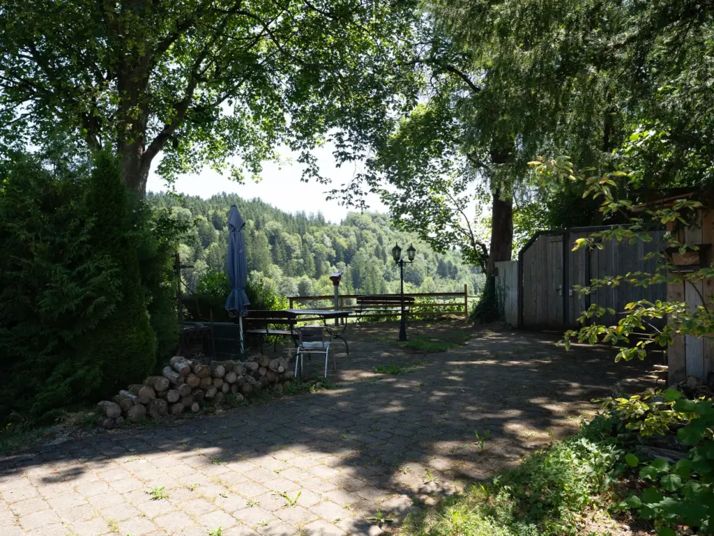 Gepflasterter Sitzplatz im Schatten großer Bäume mit Holzstapel