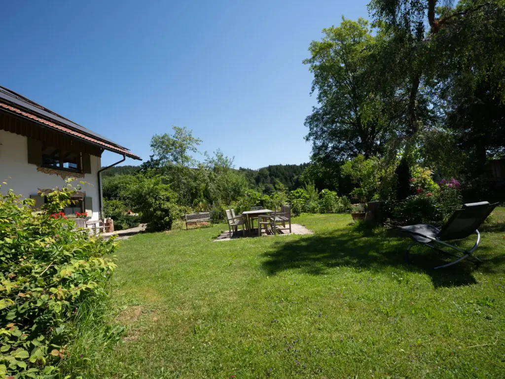 Gepflegte Wiese mit Liegestuhl vor dem Haus, dahinter Sitzgruppe und große Bäume