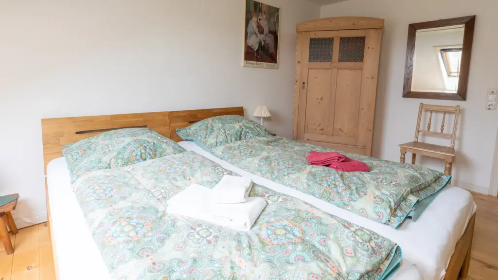 Zimmer 5: Doppelbett mit grün gemusterter Bettwäsche, Holzschrank und Spiegel unter dem Dach